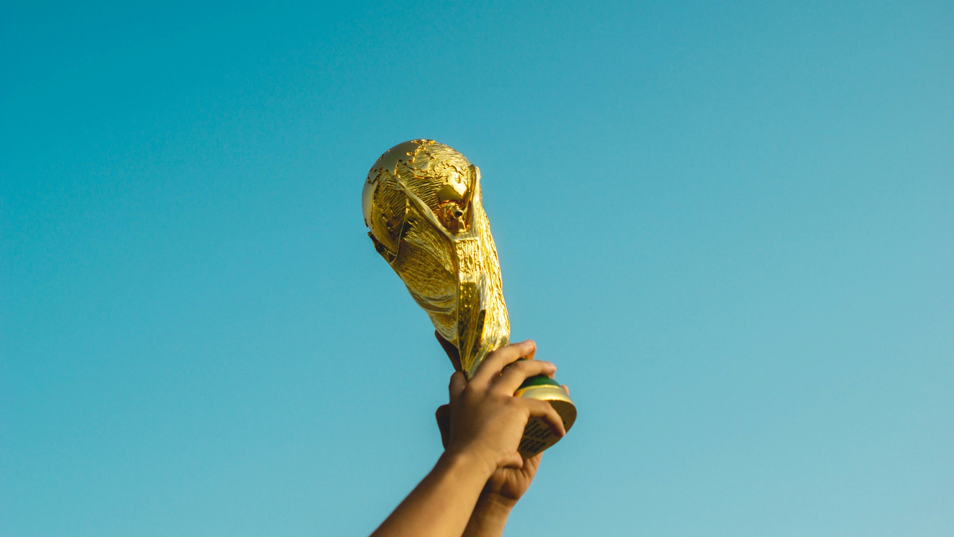 Hand holding the FIFA World Cup trophy aloft against a blue sky